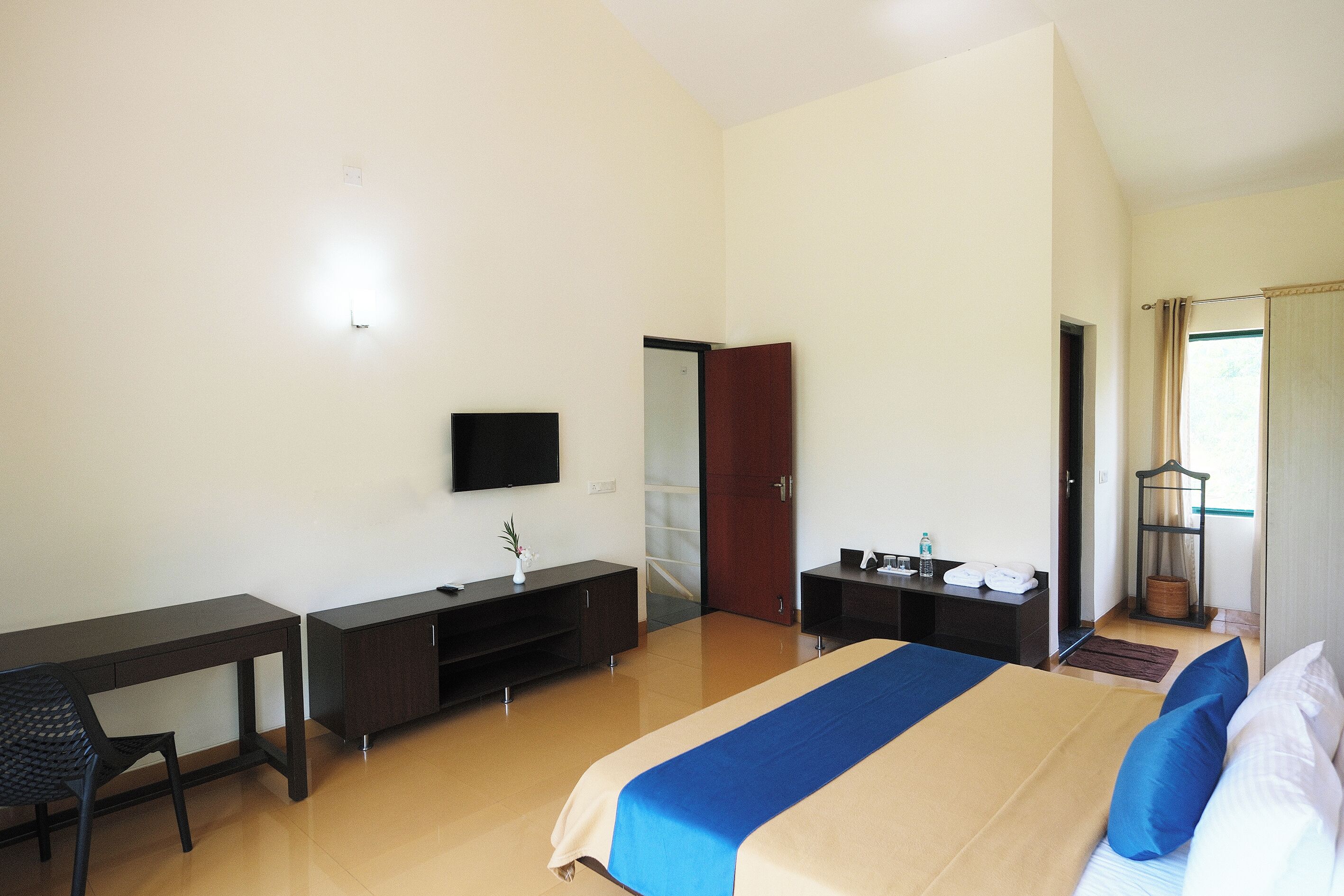 A hotel room with a bed, desk, chair, TV, wardrobe, and a small table with a lamp.