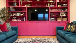 Una sala de estar con un mueble rosa para equipo electrónico, dos sofás azules, una alfombra colorida y un televisor de pantalla plana.
