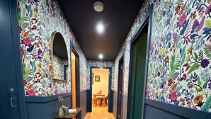 Un pasillo estrecho con suelo de madera, bordeado por paredes con papel tapiz floral, que conduce a una habitación con una mesa y sillas.