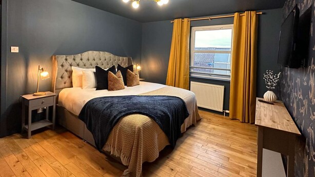 Un dormitorio con cabecera capitonada, cama con almohadas estampadas, lámpara de noche, ventana con cortinas y televisor de pantalla plana en la pared.
