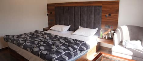 A hotel room with a large bed, a wooden headboard, a bedside table with a lamp, a chair, and a small table.
