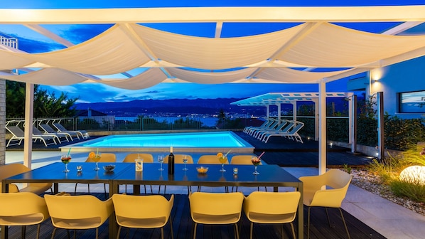 An outdoor pool area with a table and chairs, a covered seating area, and a view of the city and mountains.
