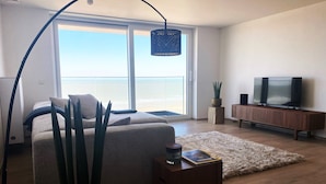 Un salon moderne avec un canapé, une table basse, un téléviseur posé sur un meuble en bois et une grande fenêtre donnant sur la mer.