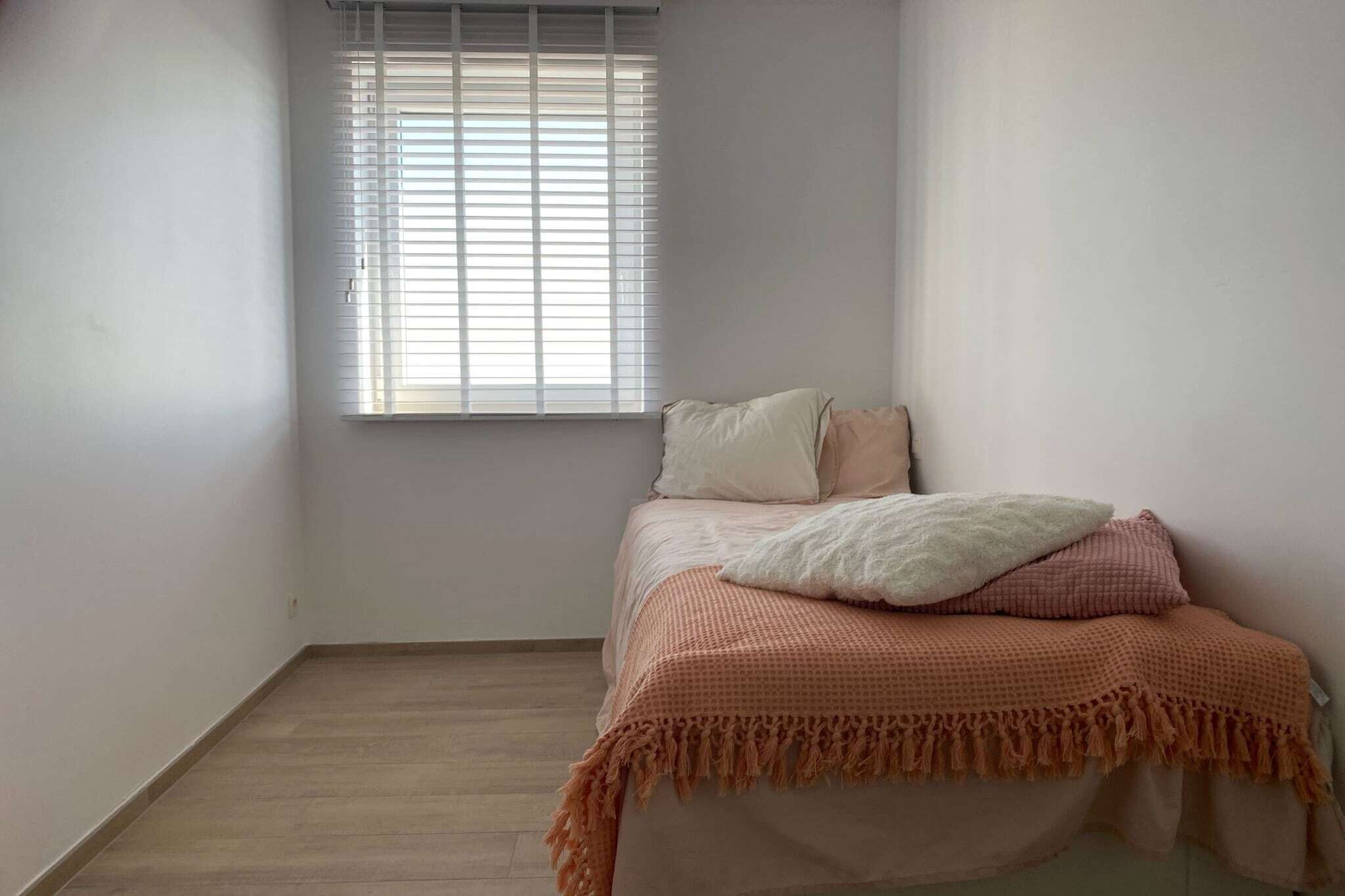 Een slaapkamer met een bed, een raam met zonwering en een houten vloer.