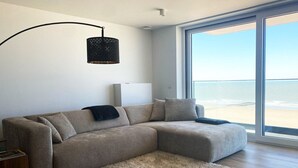 Un salon moderne avec un grand canapé d’angle, un tapis moelleux, un meuble blanc et une grande fenêtre donnant sur la plage.