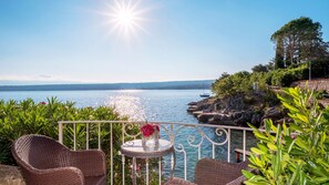 Un balcon avec des fauteuils en osier, une petite table ornée d’un vase de fleurs et une vue sur un lac où naviguent des bateaux.