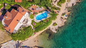 Vue aérienne d’un complexe hôtelier comprenant une piscine, une plage et un bâtiment surmonté d’un toit recouvert de tuiles rouges.