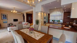 A modern living room with a wooden dining table, a sofa set, a chandelier, and a kitchen area with a window.