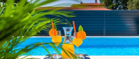A poolside setting with a table holding three glasses of orange juice, each garnished with a slice of orange and a mint leaf.