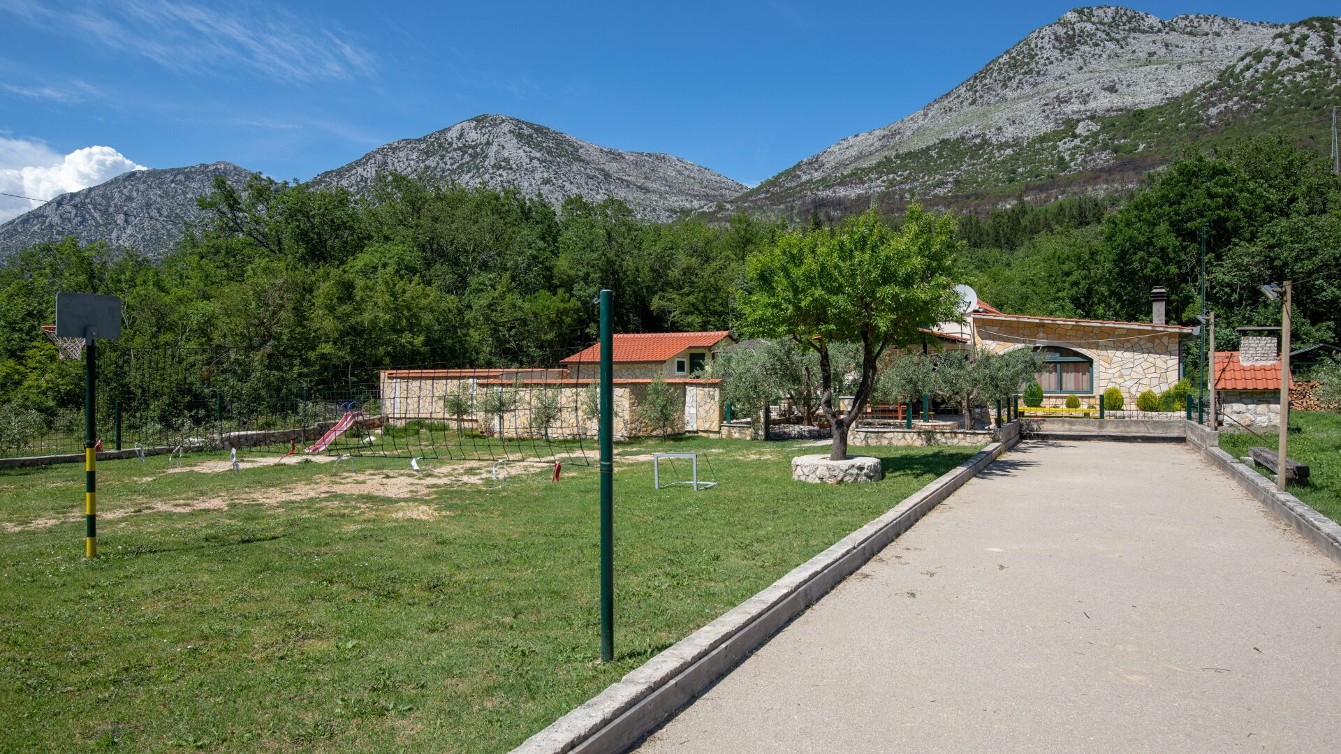 Un camí pavimentat que condueix a un edifici de pedra amb teules vermelles, envoltat de vegetació i muntanyes.