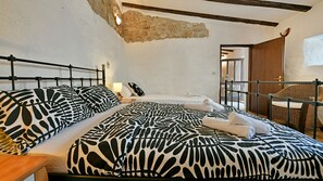 A bedroom with a bed, a nightstand, a lamp, a wicker chair, and a stone wall.