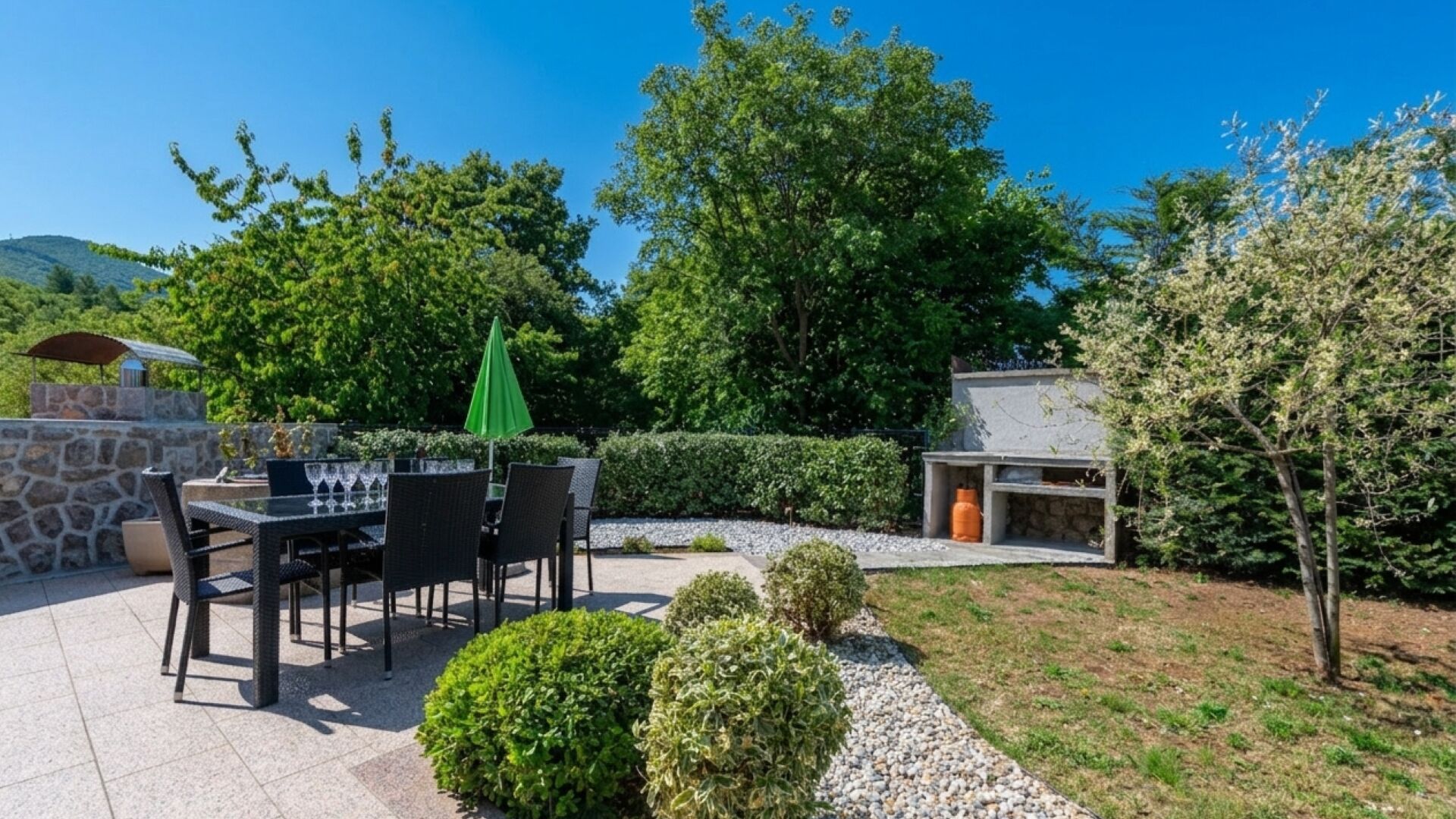 An outdoor patio with a table and chairs, a green umbrella, and a stone wall.