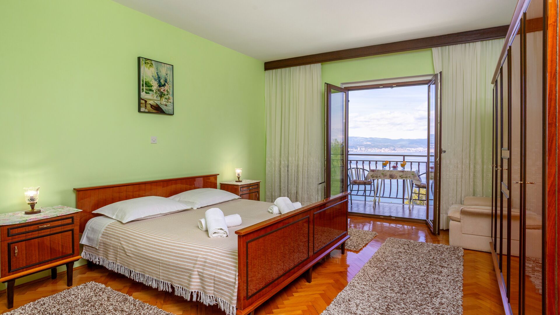 A bedroom with a bed, bedside tables, a painting, a balcony with a table and chairs, and a view of the city.