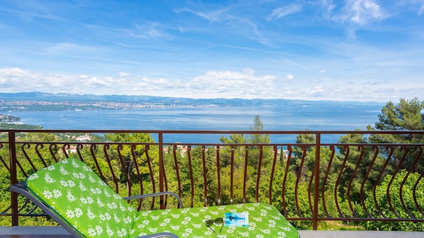 A balcony with a green floral chair, a table with a book and sunglasses, and a view of a body of water and distant land.