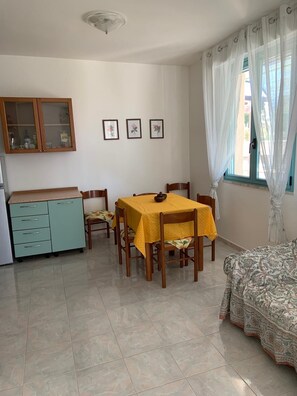 Un comedor con una mesa preparada para cuatro personas, una kitchenette con un armario y una cama con cubrecolcha estampado.