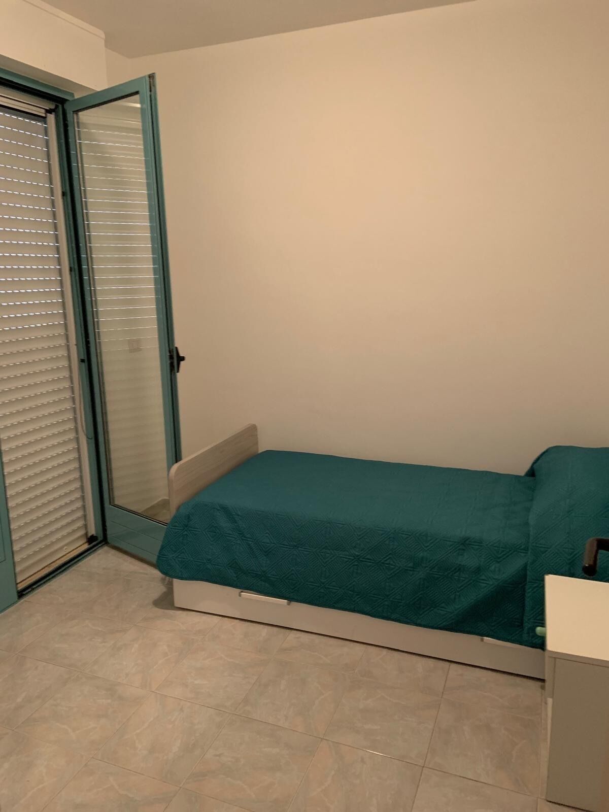 Een kleine kamer met een eenpersoonsbed, een turquoise bedovertrek, een schuifpui en een betegelde vloer.