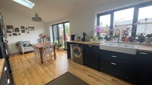 A modern kitchen with wooden flooring, a dining area with a table and chairs, and a large window with plants. - 3 Bedroom Home at Hove with Garden & Cats (Brighton and Hove)