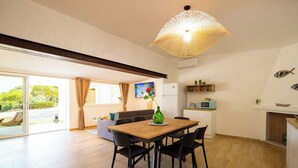 Amplia sala de estar con mesa de comedor de madera, sillas negras, una lámpara de fibra natural grande, sofá, televisor y vista a la vegetación a través de una puerta corrediza de vidrio.