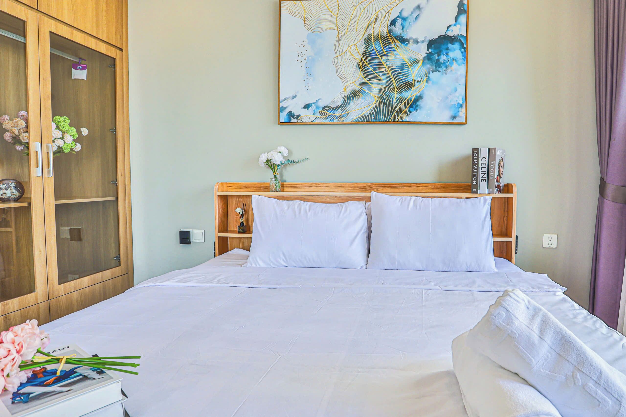 Una cama bien hecha con sábanas blancas, un cabecero de madera, un jarrón con flores y una estantería con libros.