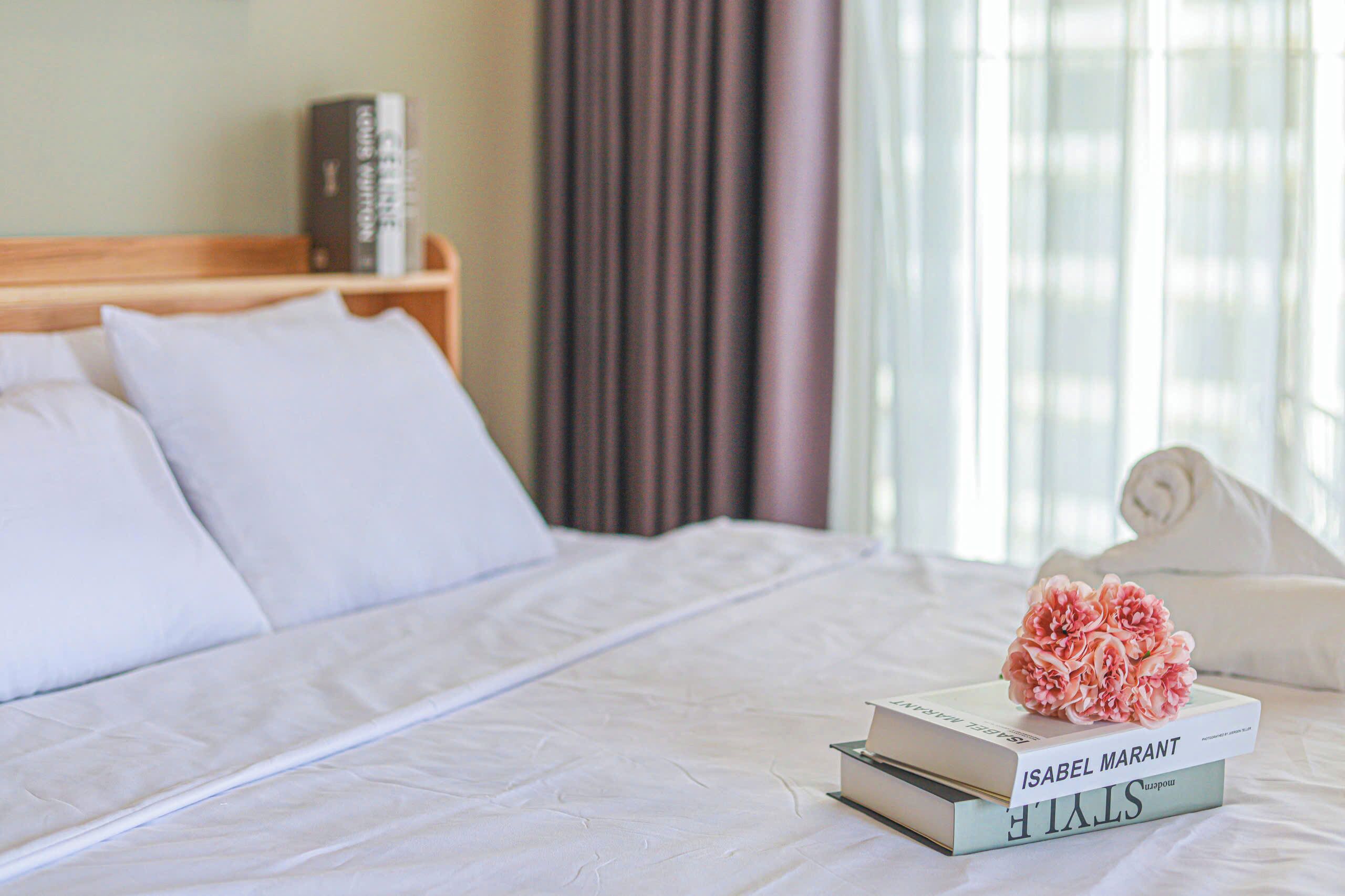 A neatly made bed with white linens, a stack of books, and a decorative pink flower on the pillows.