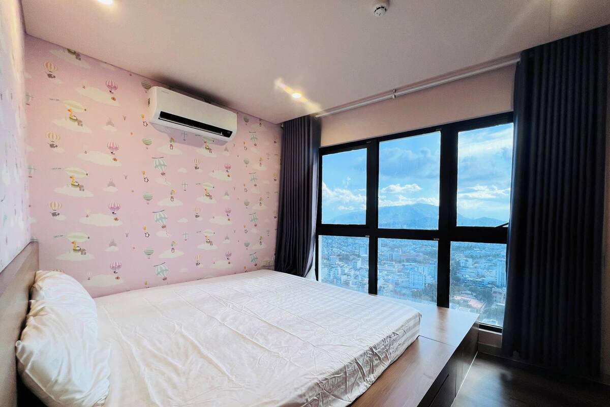 Une chambre avec un grand lit, une fenêtre donnant sur la ville et un mur rose orné d’un motif.