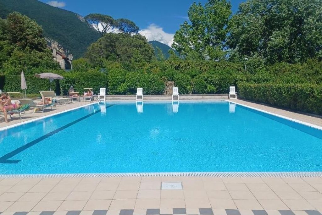 A rectangular swimming pool with clear blue water, surrounded by a tiled deck. There are lounge chairs and umbrellas along the poolside.