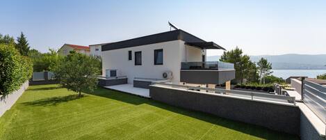 A modern two-story house with a flat roof, a balcony, and a view of the sea.