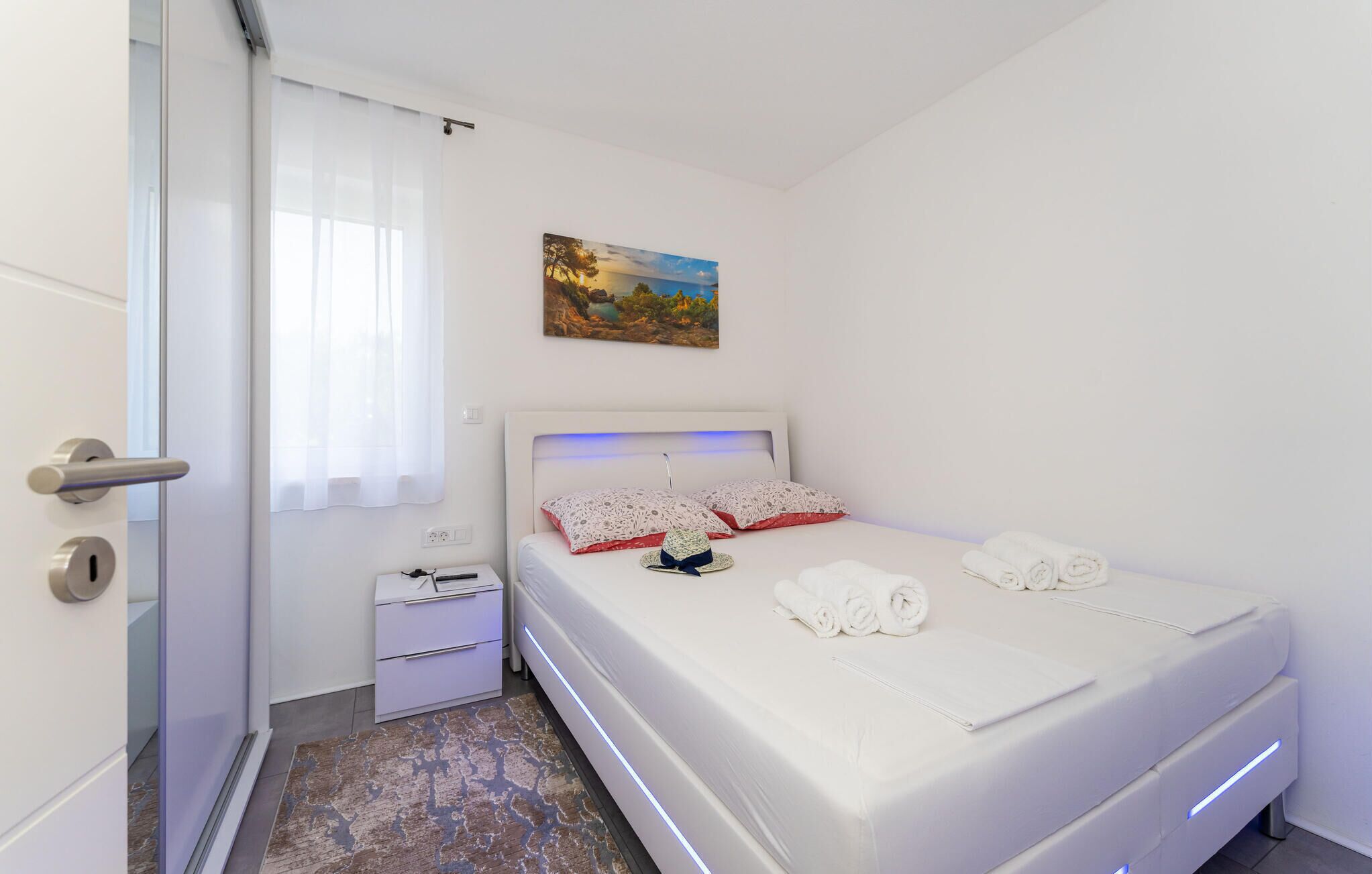 Ein modernes Schlafzimmer mit einem weißen Bett, einem Nachttisch, einem Bild an der Wand und einer Tür mit Griff.