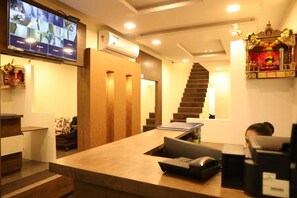 A hotel reception area with a reception desk, a staircase, and a television displaying a video call. - Galaxy Inn By Glitz Hotels (Mumbai)