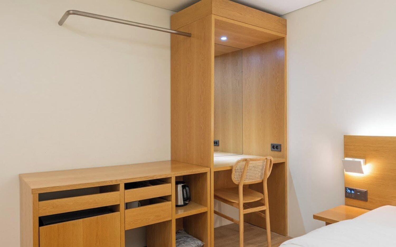 Un dormitorio pequeño con cama de madera, un escritorio y una silla. Hay una mesita de noche con una lámpara y un estante con una tetera.