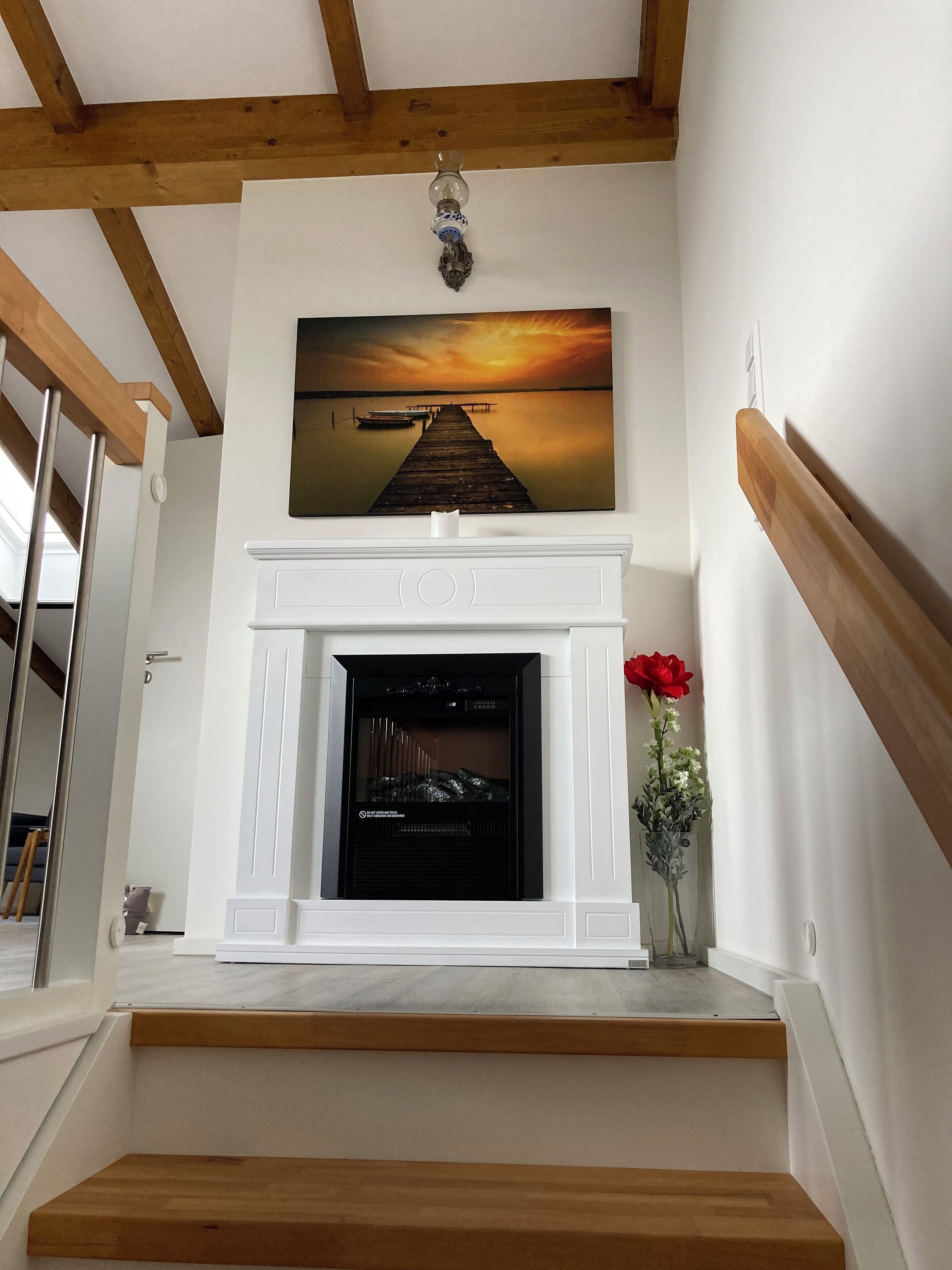 Een open haard met daarboven een schilderij, een rozenvaas en houten balken in het plafond.