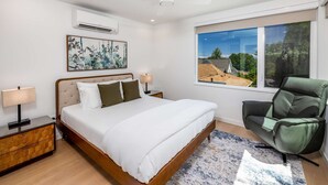 A bedroom with a bed, a chair, a nightstand, a lamp, a ceiling fan, and a window with a view of buildings and trees. - Secret Hideaway - New Custom Built Home (Seattle)