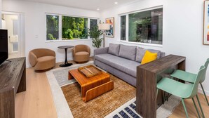A modern living room with a grey sofa, two armchairs, a coffee table, and a TV. Large windows offer views of greenery outside. - Secret Hideaway - New Custom Built Home (Seattle)