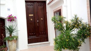 Une entrée d’immeuble avec une double porte en bois, des plantes en pot et un mur blanc portant un numéro.