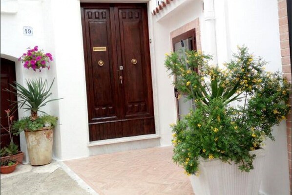 A building entrance with a double wooden door, potted plants, and a white wall with a number sign.