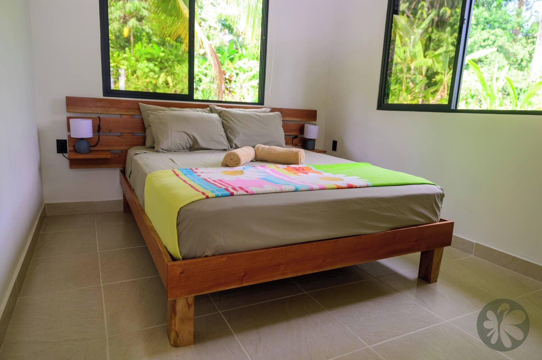 Un letto con struttura in legno, biancheria beige e una coperta patchwork colorata. Due lampade da parete sono poste ai lati del letto.
