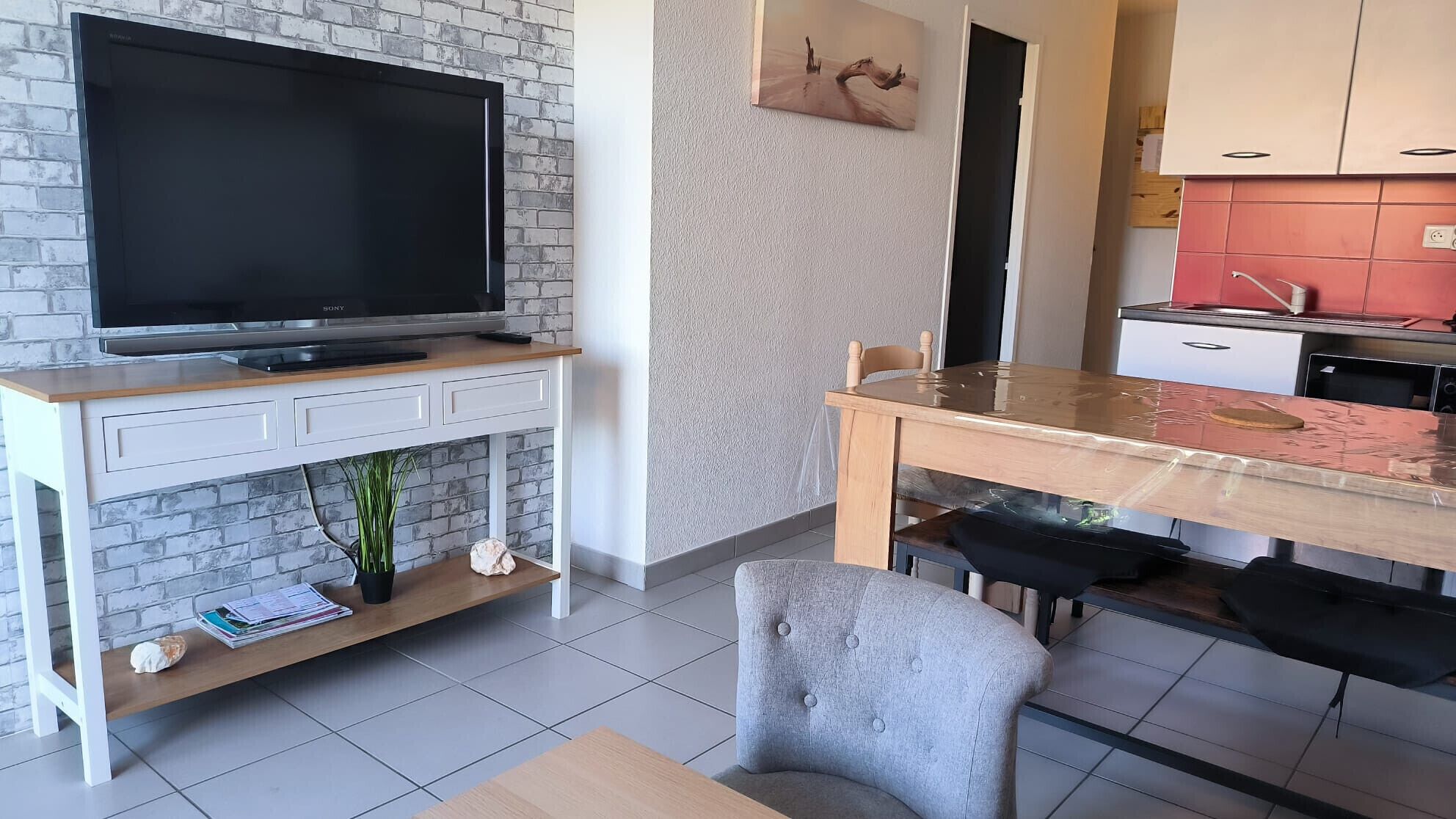 A modern living space with a flat-screen TV on a white console table, a dining area with a wooden table and chairs, and a kitchen with red cabinets.