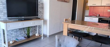 A modern living space with a flat-screen TV on a white console table, a dining area with a wooden table and chairs, and a kitchen with red cabinets.