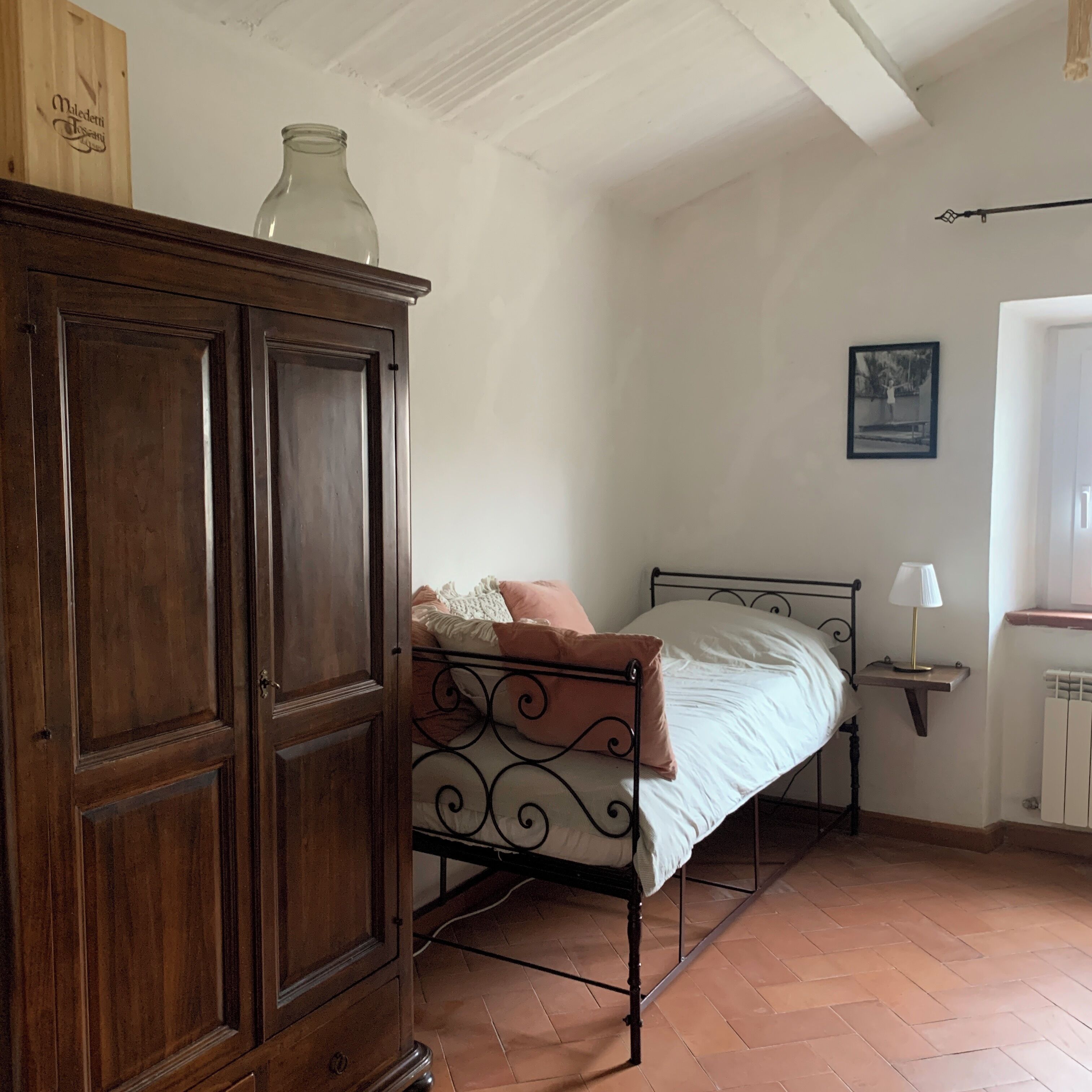 A bedroom with a wooden wardrobe, a metal bed frame, a bedside table with a lamp, and a radiator.