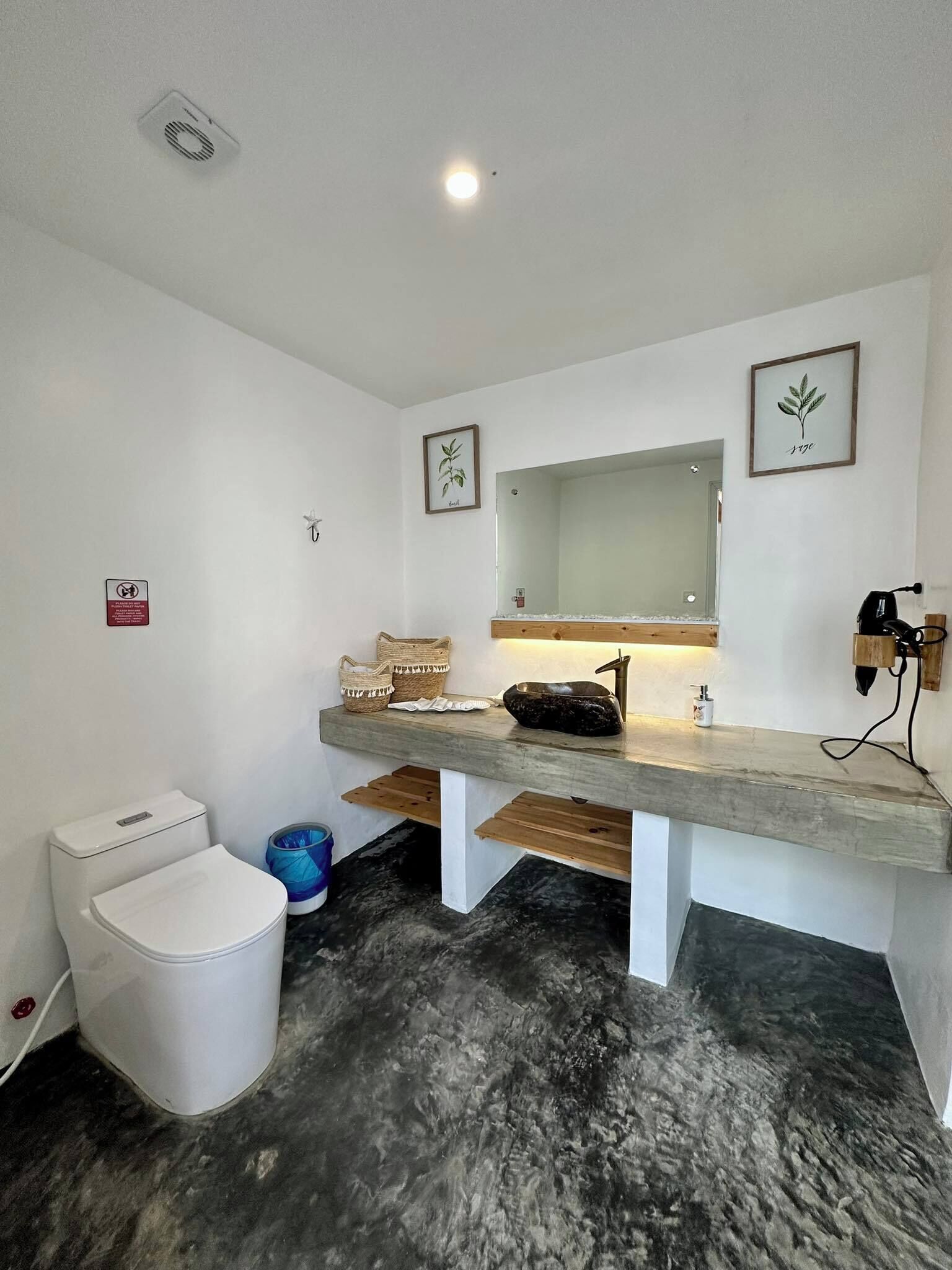 Un baño moderno con inodoro blanco, lavamanos con encimera de madera y espejo encima. Hay impresiones botánicas enmarcadas en la pared y un pequeño cubo de basura junto al inodoro.