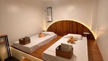A hotel room with two single beds, a ceiling fan, an air conditioning unit, a small table, and a bench.