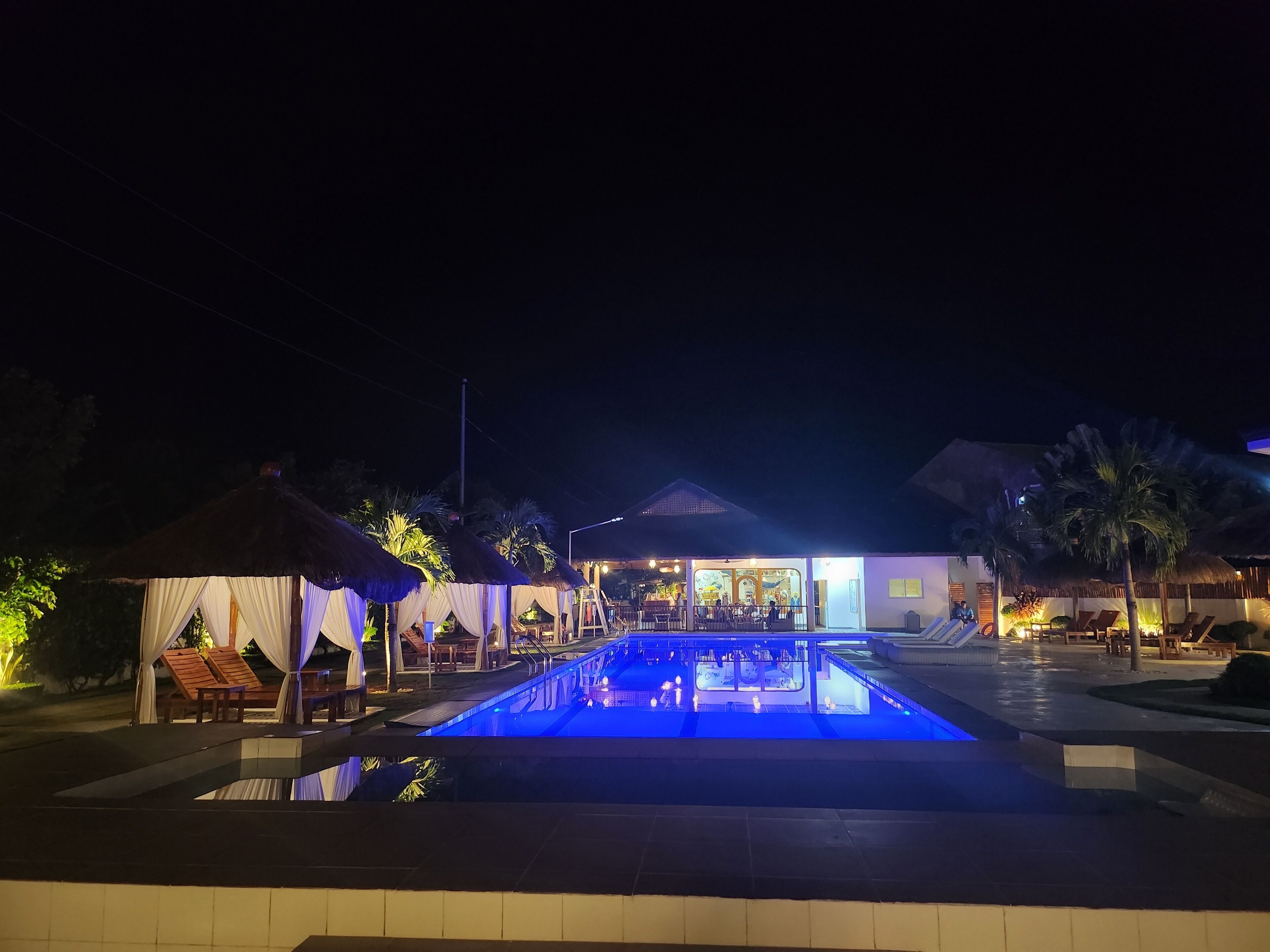 A nighttime view of a resort with a swimming pool, lounge chairs, and thatched-roof structures.