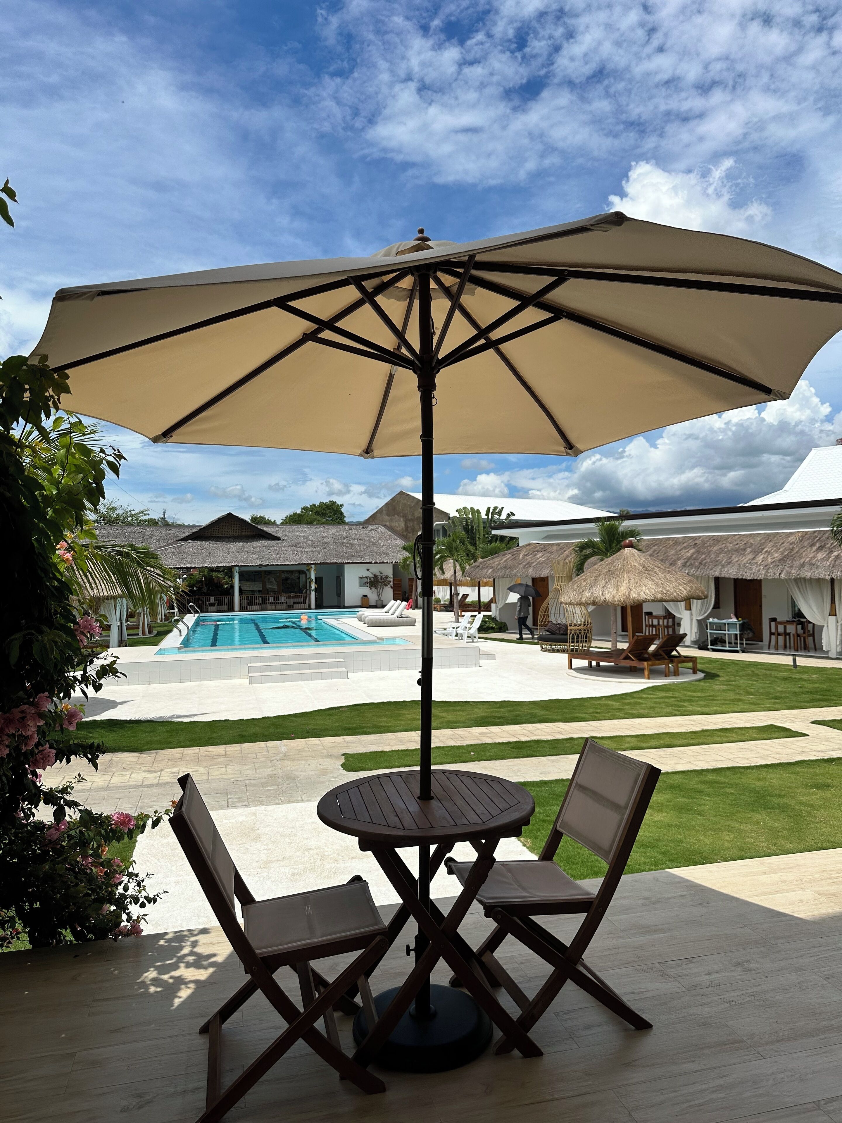 Une terrasse avec une table et des chaises, protégée par un grand parasol, d’où l’on peut admirer une piscine et des bâtiments aux toits de chaume.