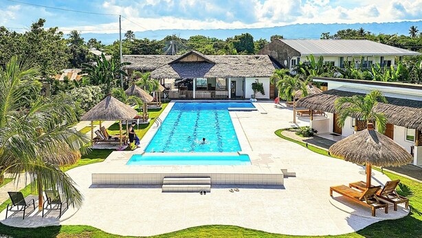 A swimming pool surrounded by thatched-roof structures, a grassy area, and a view of distant mountains.