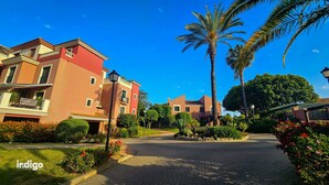 A residential area with a paved driveway, palm trees, and a building with balconies. - Apartment 2 bedrooms • Pool View • Prado Golf • DAR01 (Isla Canela)
