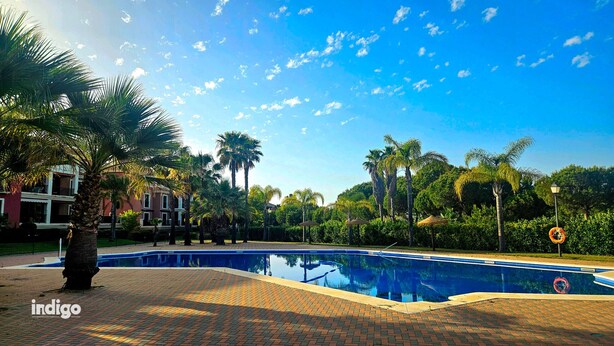 A swimming pool with a brick pathway, palm trees, and a building in the background. - Apartment 2 bedrooms • Pool View • Prado Golf • DAR01 (Isla Canela)