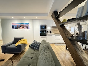 Ein modernes Wohnzimmer mit einem grauen Sofa, einem Holztisch und einer Küche im Hintergrund.