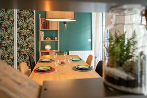 A dining area with a wooden table set for four, surrounded by chairs. There is a bookshelf with books and decorative items, a lamp, and a glass container with plants.