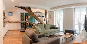 A modern living room with a green sofa, a grey armchair, and a wooden coffee table. A staircase with a wooden railing leads to an upper level.