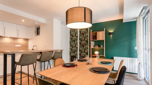 A modern kitchen with a wooden table set for dining, a hanging light fixture, and a green accent wall. - Charming apartment 5P (Cauterets)